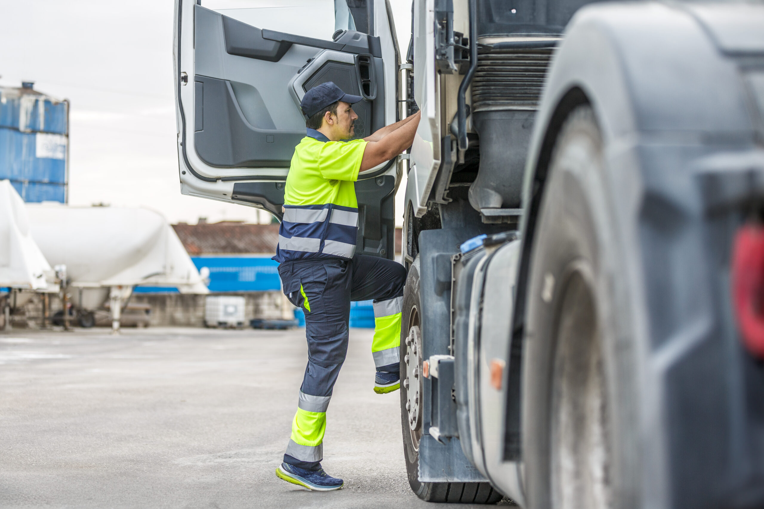 Full body side view of focused male driver in uniform and cap climbing steps while entering truck to commence factory delivery operation