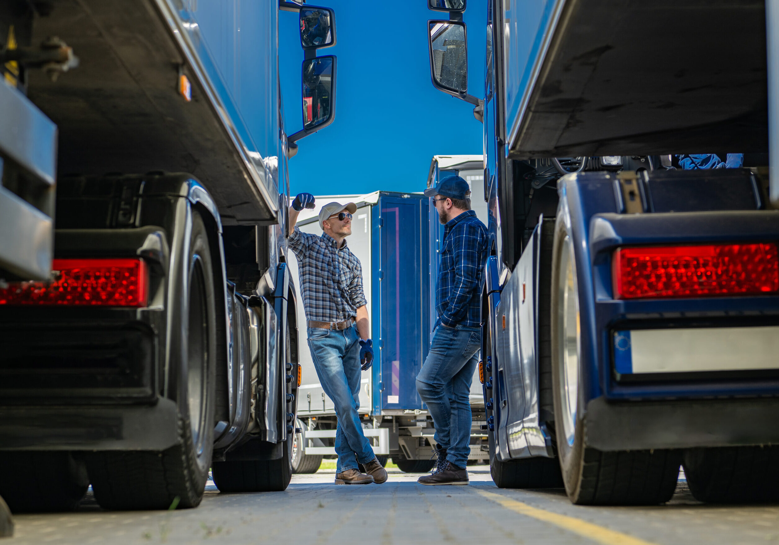 Two truckers are standing in front of a large semi truck. One man is holding a clipboard and the other is looking at the trucks engine. The truck is parked in a paved lot with other vehicles in the background.